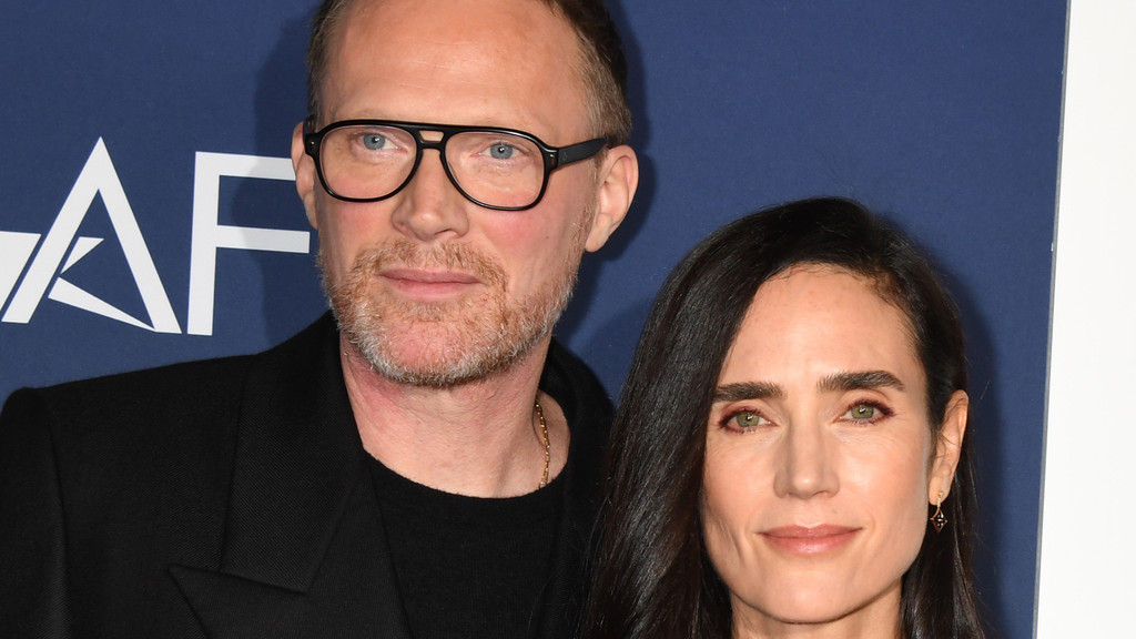 Paul Bettany, wearing glasses and a beard, and Jennifer Connelly, with dark hair and green eyes, stand close together, looking forward, against a blue background with a white "AF" logo.