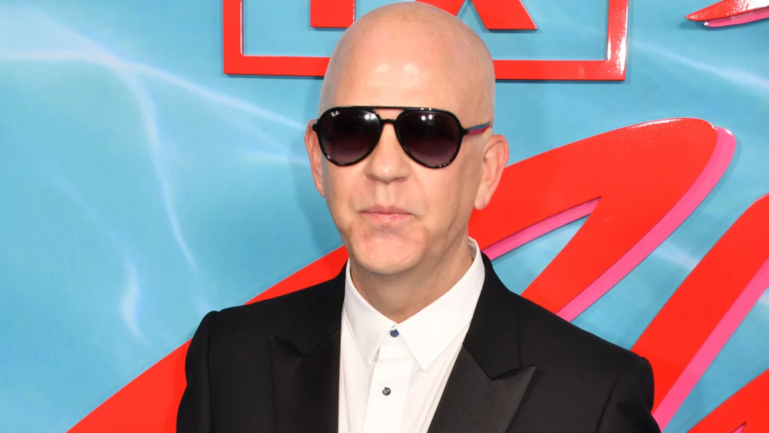 A bald man wearing dark Ray-Ban sunglasses, a black suit, and a white shirt stands against a light blue background with abstract red and pink designs.