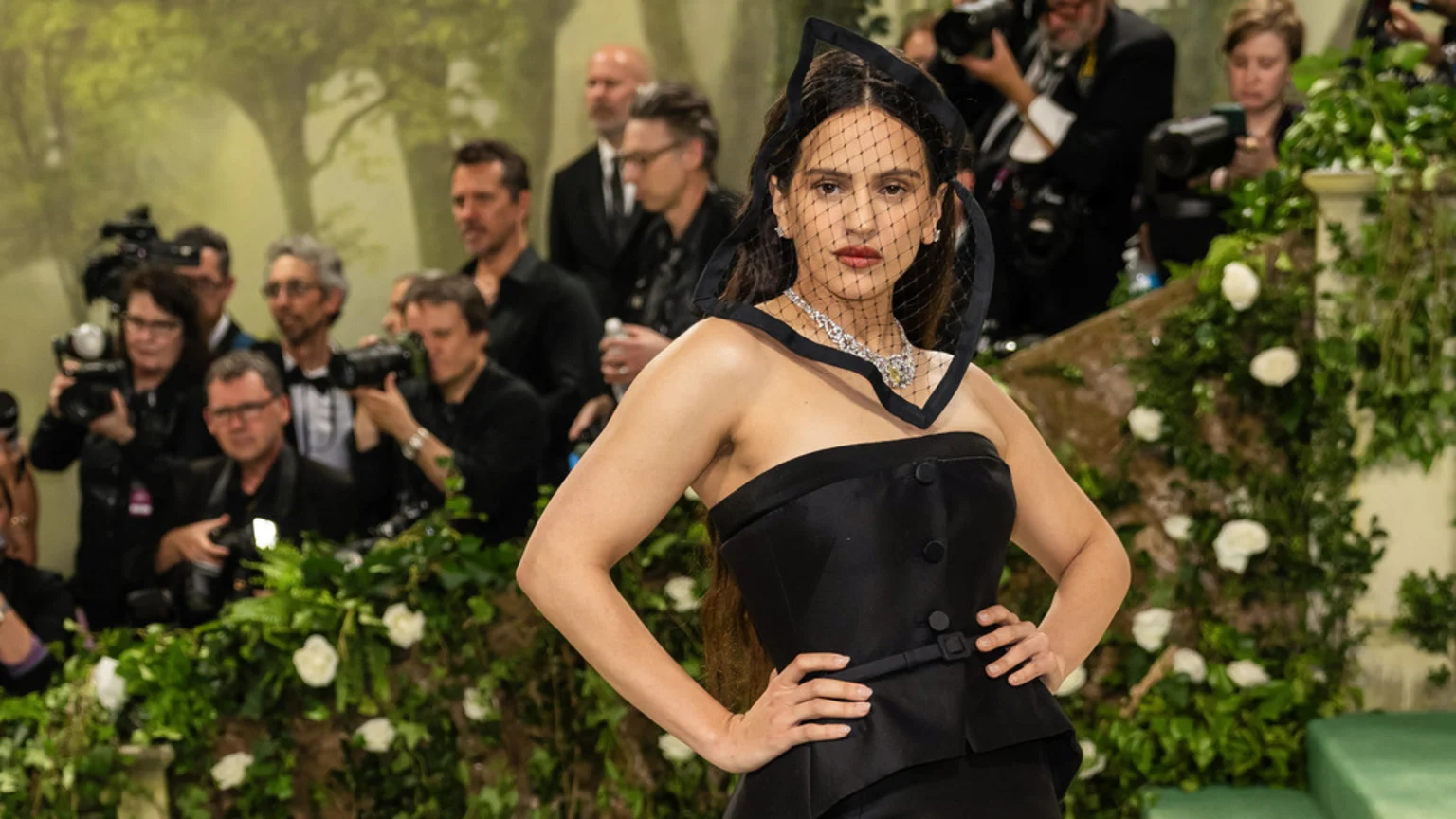 Rosalía, wearing a black strapless top, a veiled headpiece, and a diamond necklace, poses with her hands on her hips for photographers in a lush garden setting.
