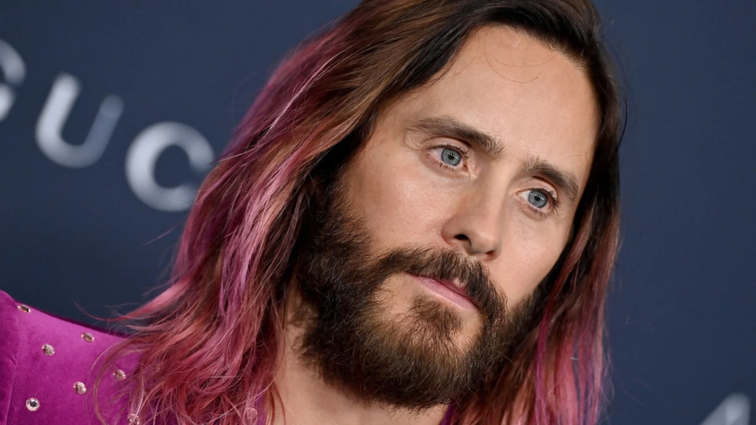Close-up of actor Jared Leto with long, pink-tinted hair and a beard, wearing a magenta top, looking off-camera against a blurry dark background with a "GUCCI" logo.