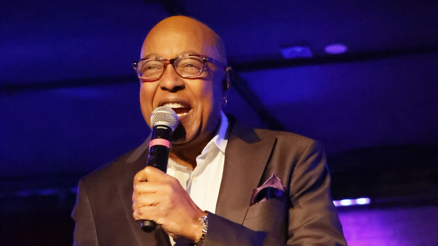 A bald Black man wearing glasses and a suit with a purple pocket square sings into a Shure microphone on a stage lit with blue and purple lights, his mouth open in song.