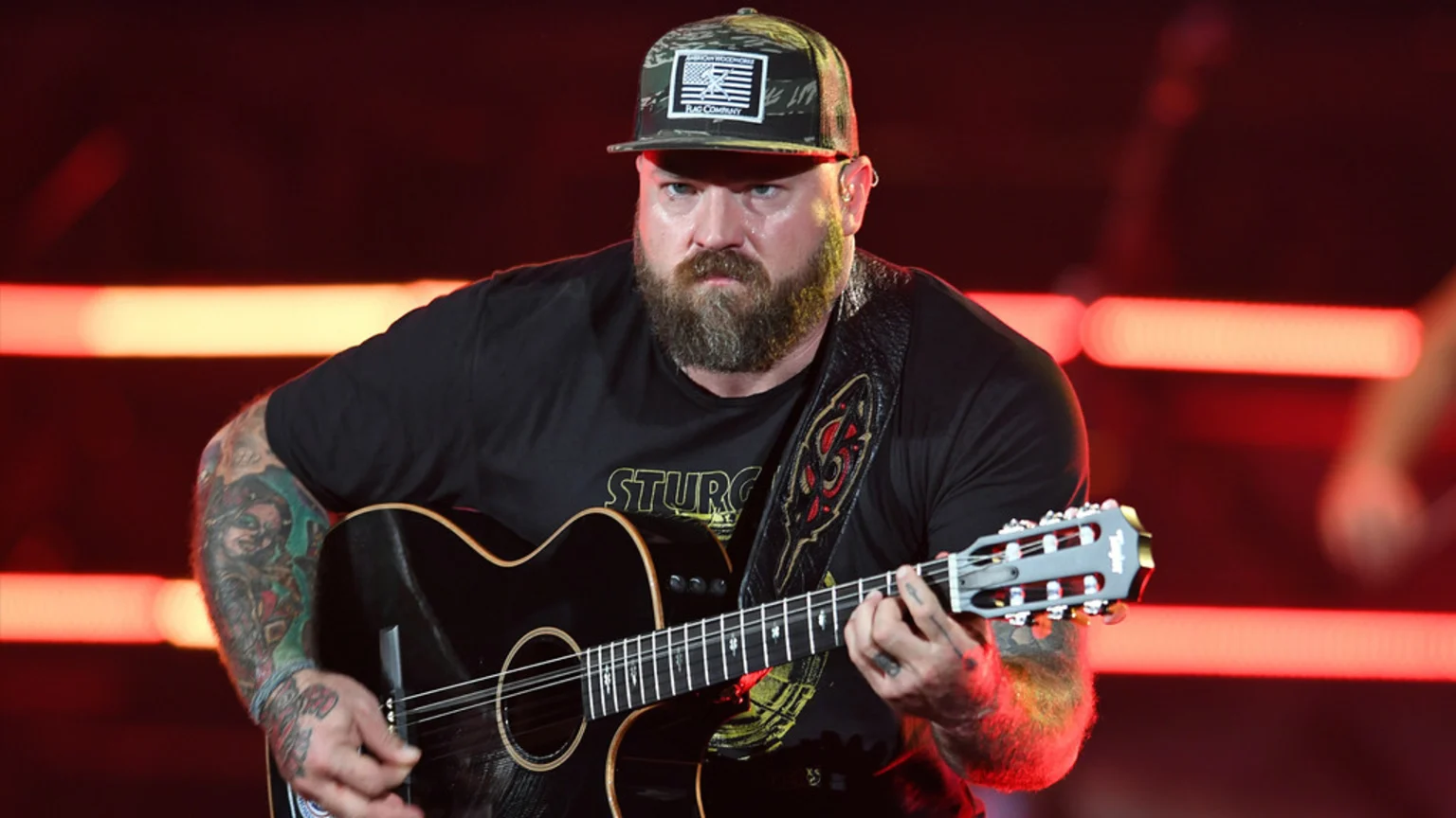 Zac Brown, a bearded man in a camouflage cap and black t-shirt, plays a dark acoustic guitar on stage, his tattooed arms visible against a red-lit background.