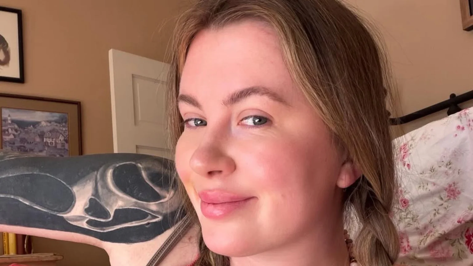 A woman with light brown hair in pigtails smiles at the camera, revealing a large black and white tattoo on her arm, with framed artwork and a floral bedsheet in the background.