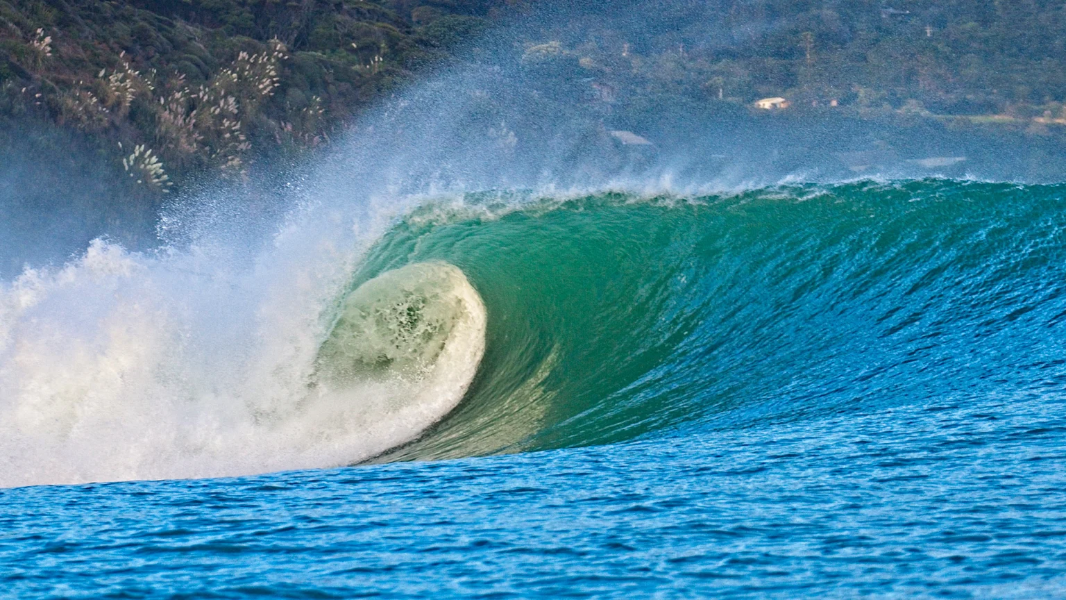 A powerful blue-green ocean wave with a curling barrel, generating white spray, against a backdrop of green hills.