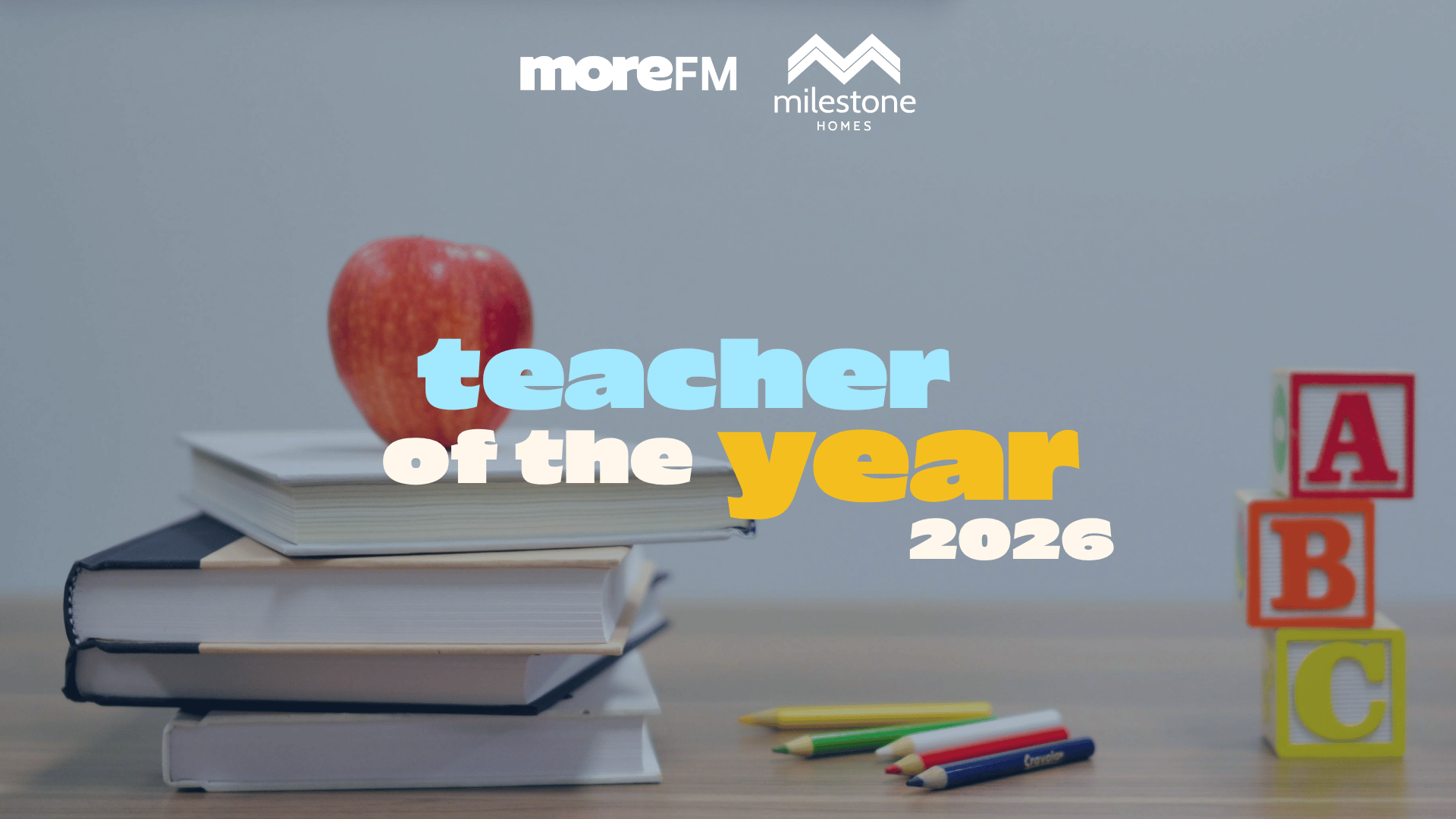 A red apple sits atop a stack of books, alongside colored pencils and ABC blocks, with "moreFM", "milestone HOMES", and "teacher of the year 2026" text overlaid.