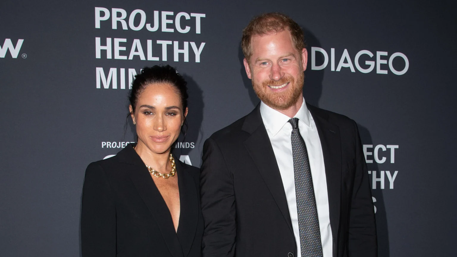 Meghan Markle in a black blazer and Prince Harry in a suit smile at the camera in front of a dark gray background with event branding.
