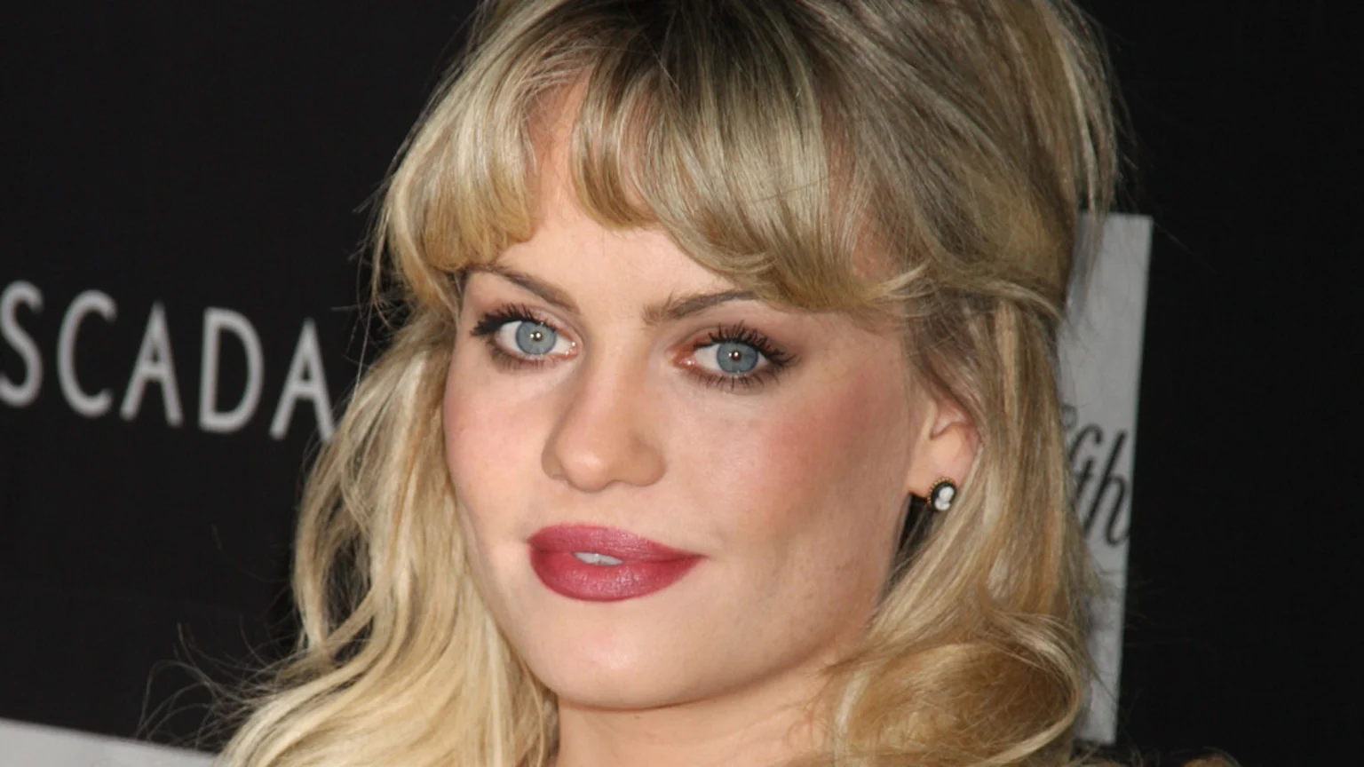 A blonde woman with blue eyes and bangs, wearing red lipstick and a cameo earring, looks directly forward against a dark background with "ESCADA" written on it.