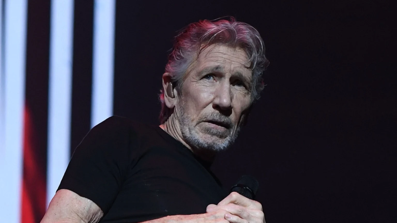 Roger Waters, an older man with graying hair and beard, wearing a black t-shirt, holds a microphone and has an in-ear monitor while on stage with vertical lights in the background.
