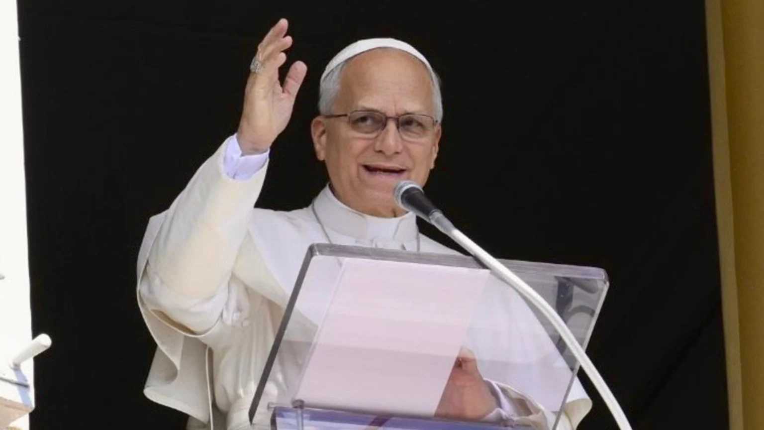 A smiling man in a white skullcap and religious attire, wearing glasses, stands behind a clear podium with a microphone, raising his right hand in a wave or blessing gesture.