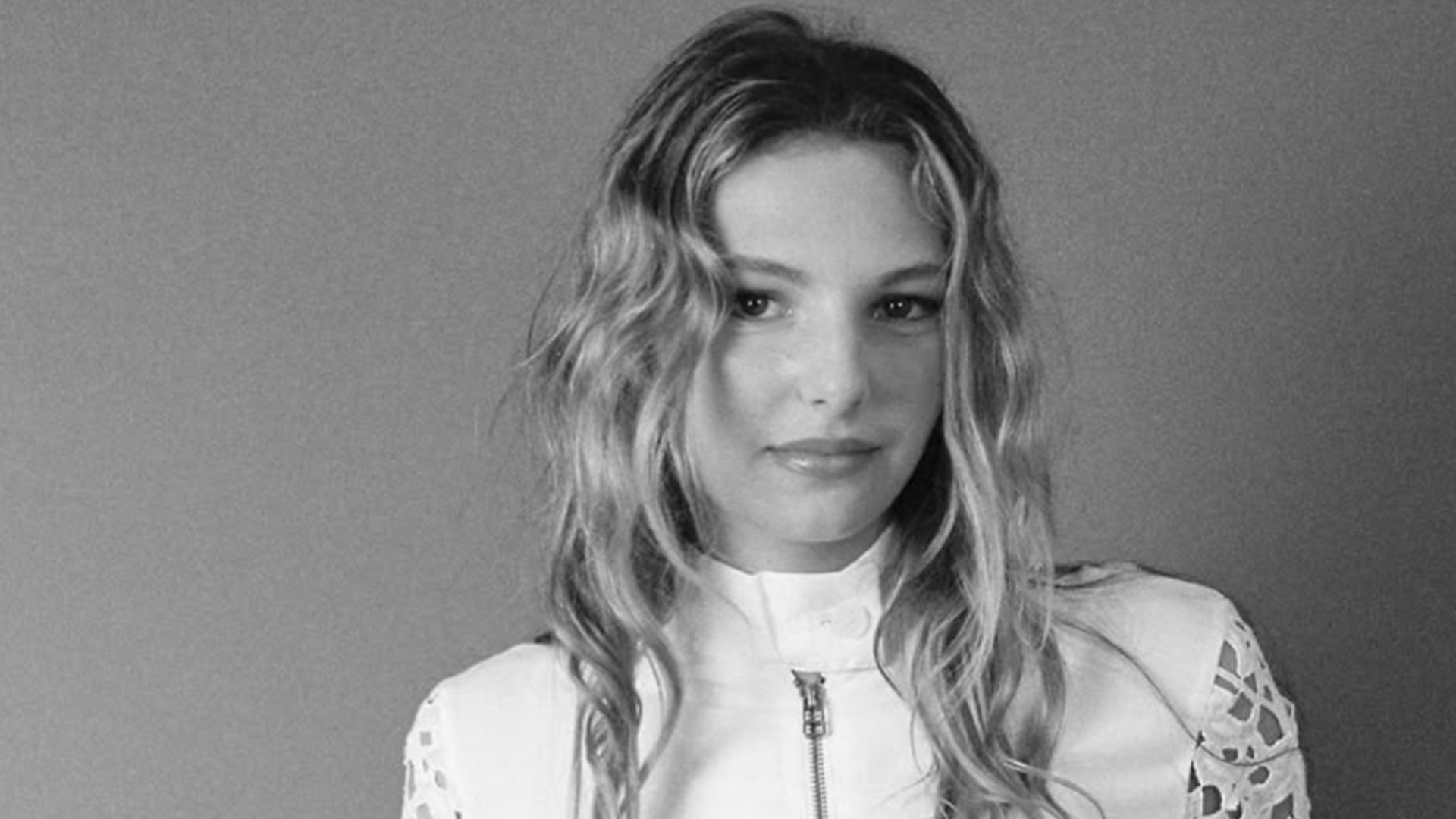 A black and white close-up shows a young woman with long wavy hair, looking at the camera with a gentle smile, wearing a white zippered top with intricate shoulder details against a plain grey background.