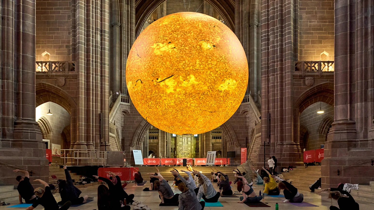 People doing yoga poses on mats beneath a large, glowing, spherical yellow-orange object resembling the sun, inside a grand stone building.