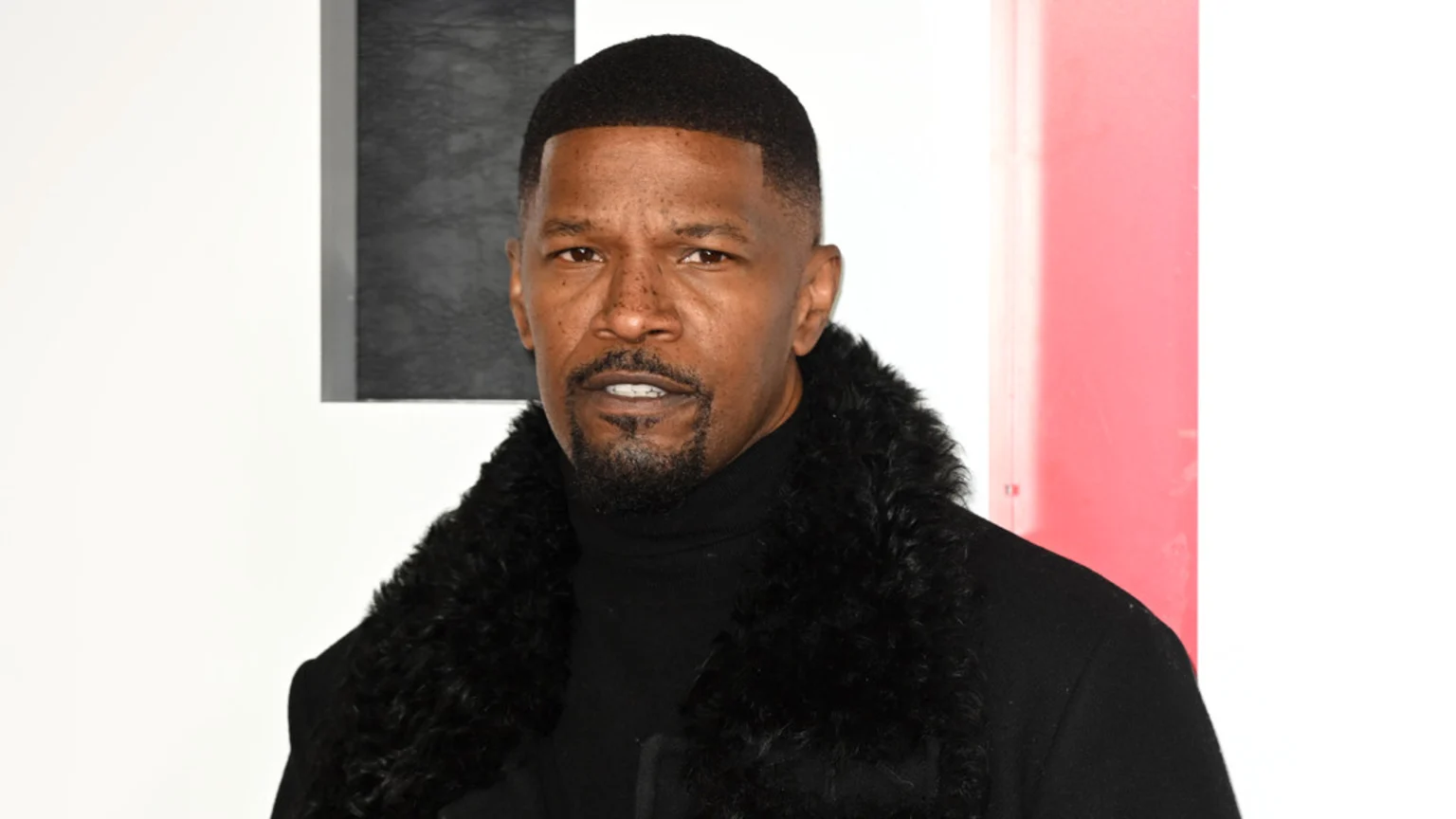 Jamie Foxx looks directly at the camera, wearing a black turtleneck and a black coat with a furry collar, against a white, grey, and red background.
