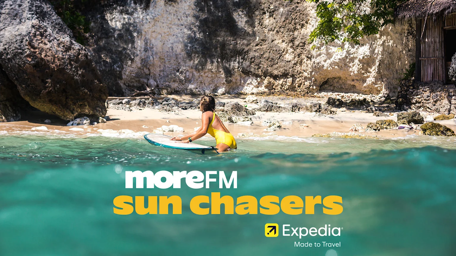 A vibrant beach scene with a woman in a yellow swimsuit pushing a surfboard into clear ocean water, backed by sandy cliffs and foliage, with "moreFM sun chasers" and "Expedia" logos.