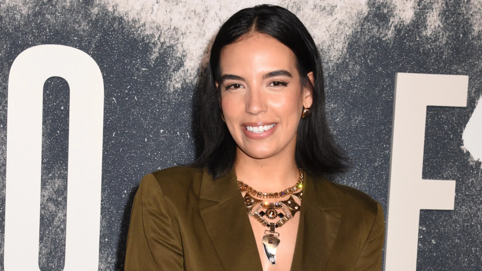 Danielle Olivera, with dark hair parted down the middle, smiles confidently at the camera, wearing a dark green blazer, gold hoop earrings, and an ornate gold statement necklace.