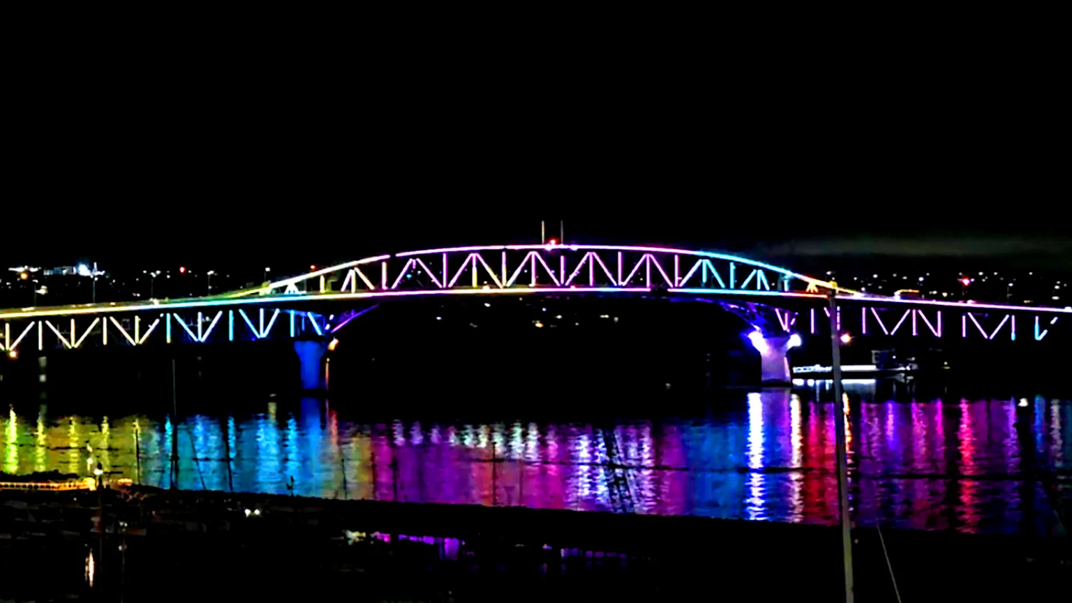 A nighttime photograph of an arch truss bridge brightly lit with rainbow colors spanning dark reflective water. The bridge's multicolored lights create vivid reflections, with a distant cityscape visible under the night sky and small boats or docks silhouetted at the water's edge.