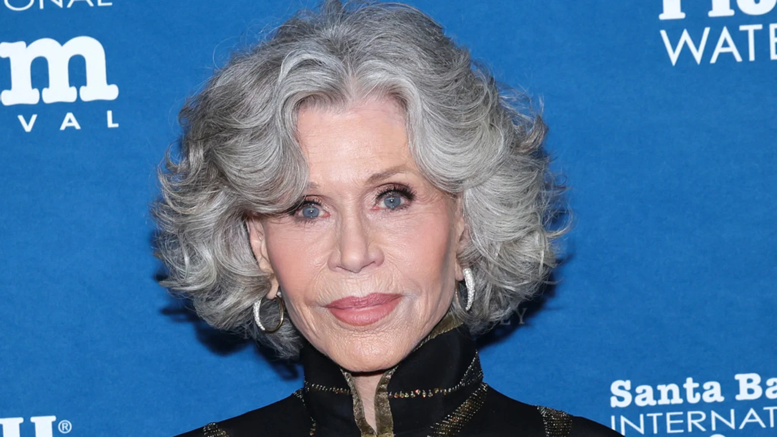 Jane Fonda, an older woman with gray, curly hair and blue eyes, smiles faintly while wearing sparkling hoop earrings and an embellished dark top against a blue event backdrop.