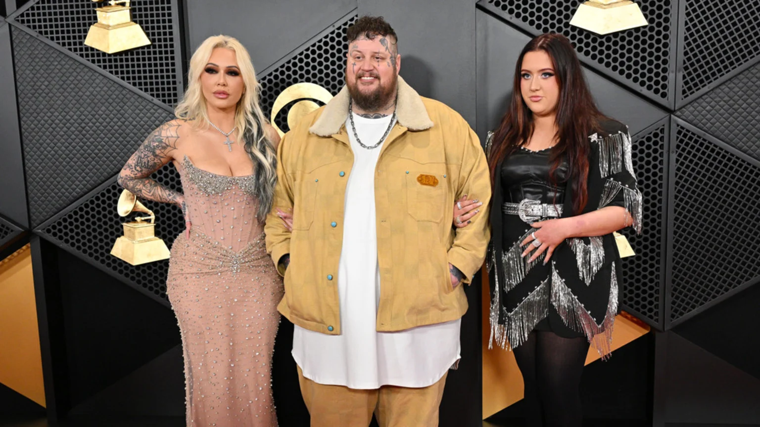 Jelly Roll, Bunnie Xo, and his daughter Bailee Ann pose on a red carpet, with Bunnie Xo in a nude embellished dress, Jelly Roll in a yellow jacket, and Bailee Ann in a black fringe dress, against a Grammy awards backdrop.