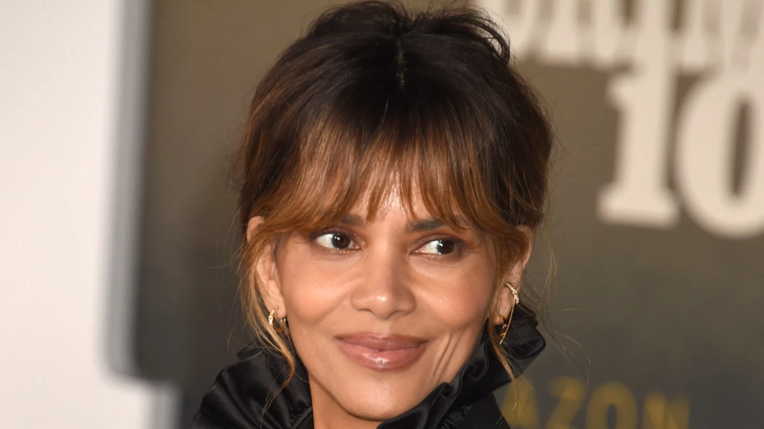 A close-up of actress Halle Berry smiling gently, with dark hair and light brown bangs, wearing a black ruffled top and earrings, looking slightly to her left.