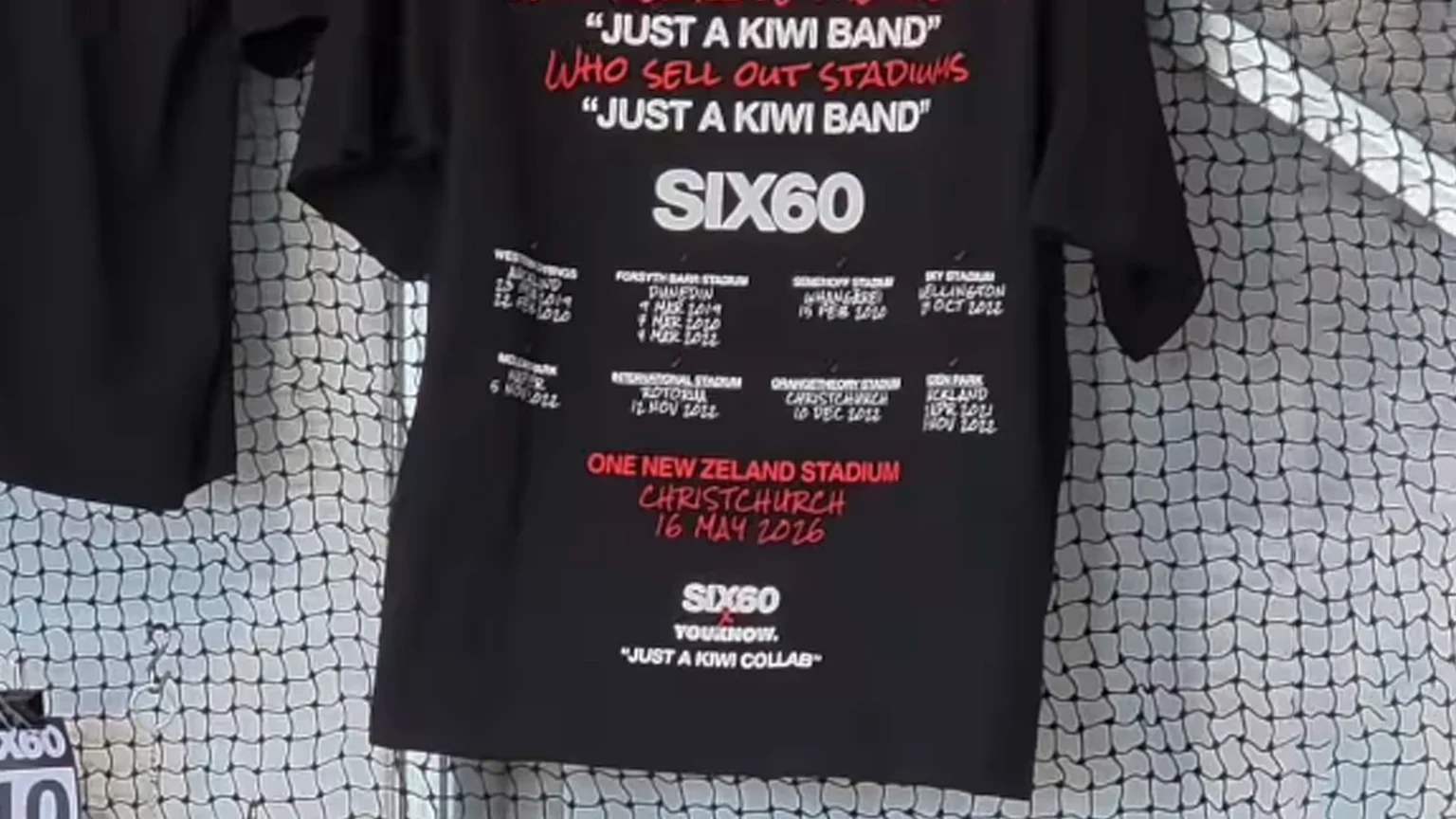 A black Six60 band t-shirt hangs on a white grid, displaying concert dates and venues, with top white and red text stating "JUST A KIWI BAND WHO SELL OUT STADIUMS", and a red section for "ONE NEW ZELAND STADIUM CHRISTCHURCH 16 MAY 2026".