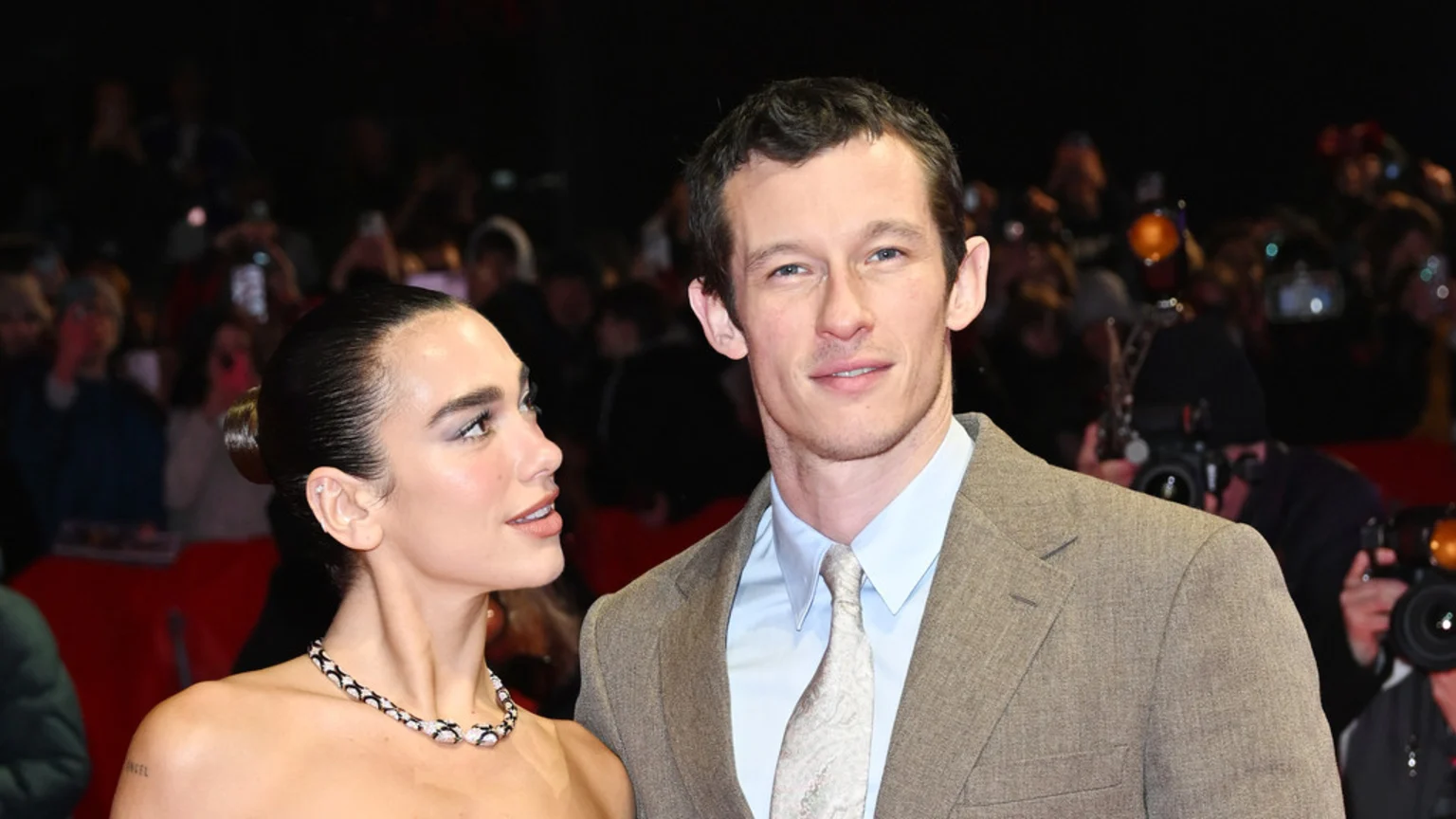 Dua Lipa, wearing a strapless top, necklace, and shoulder tattoo, stands next to a man in a suit and tie at an event, with a blurred crowd in the background.