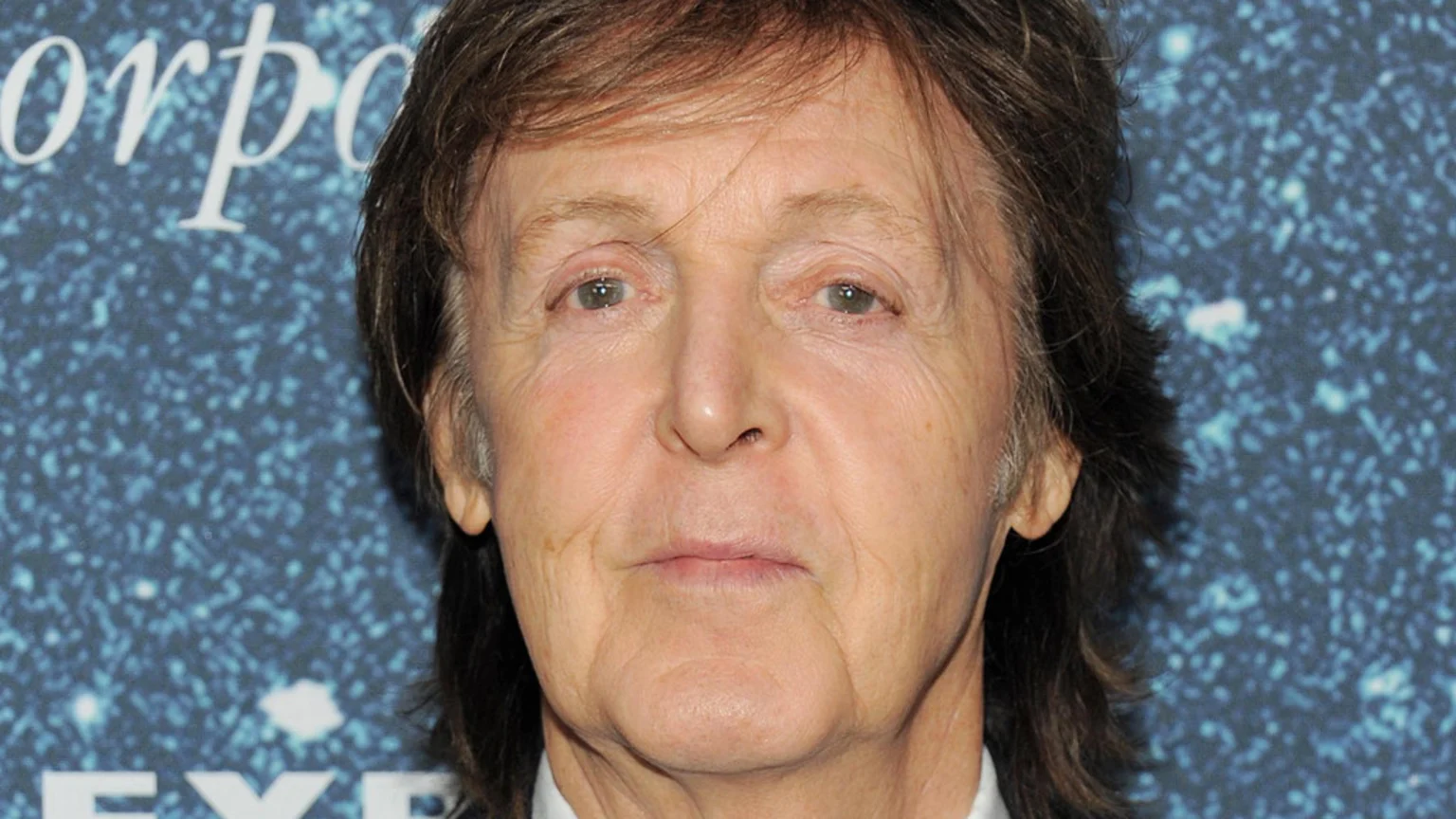A close-up headshot of an older Paul McCartney with light brown hair and a neutral expression, against a blurry dark blue background with white specks and partial white text.