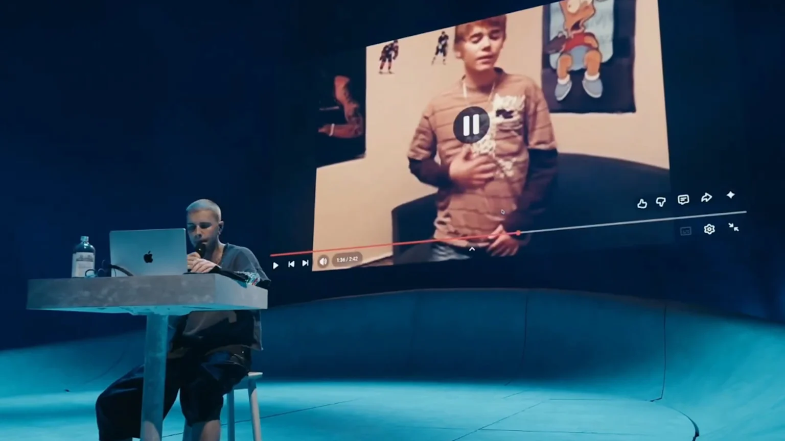 A person with short light hair sits at a desk with a laptop and microphone, watching a large screen showing a YouTube video of a young Justin Bieber, with playback controls overlaid, in a dark blue room.
