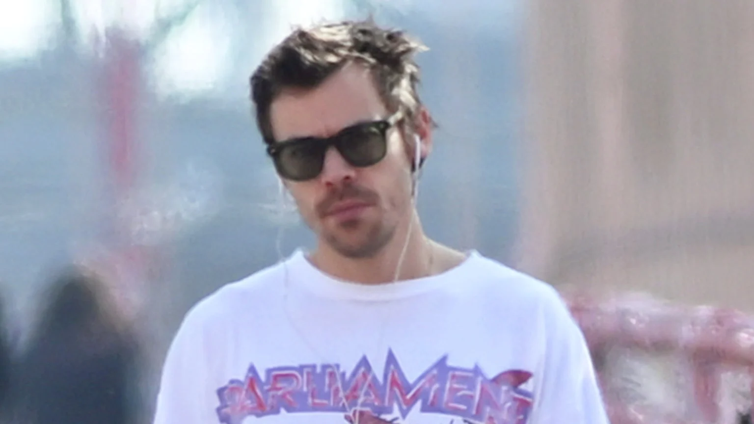 Close-up of Harry Styles wearing dark sunglasses, white wired earbuds, a mustache, and a white t-shirt with "PARLIAMENT" on it, looking forward against a blurry light background.