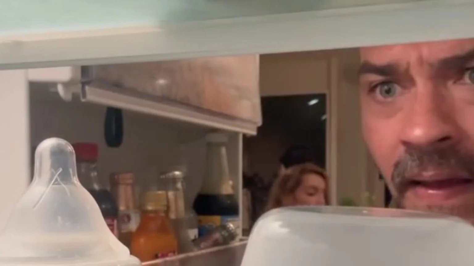 A man with wide eyes peeks into a refrigerator, where a baby bottle is prominent in the foreground among other items, and a woman is partially visible in the background.