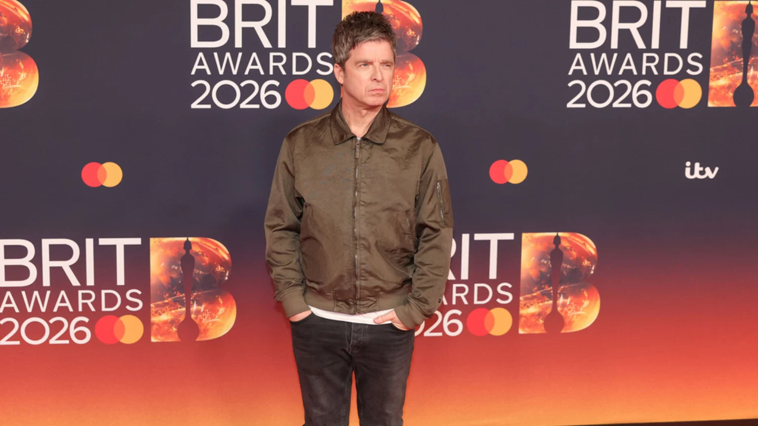 Noel Gallagher stands in front of a BRIT Awards 2026 backdrop, wearing a green bomber jacket, white t-shirt, and dark jeans with his hands in his pockets.