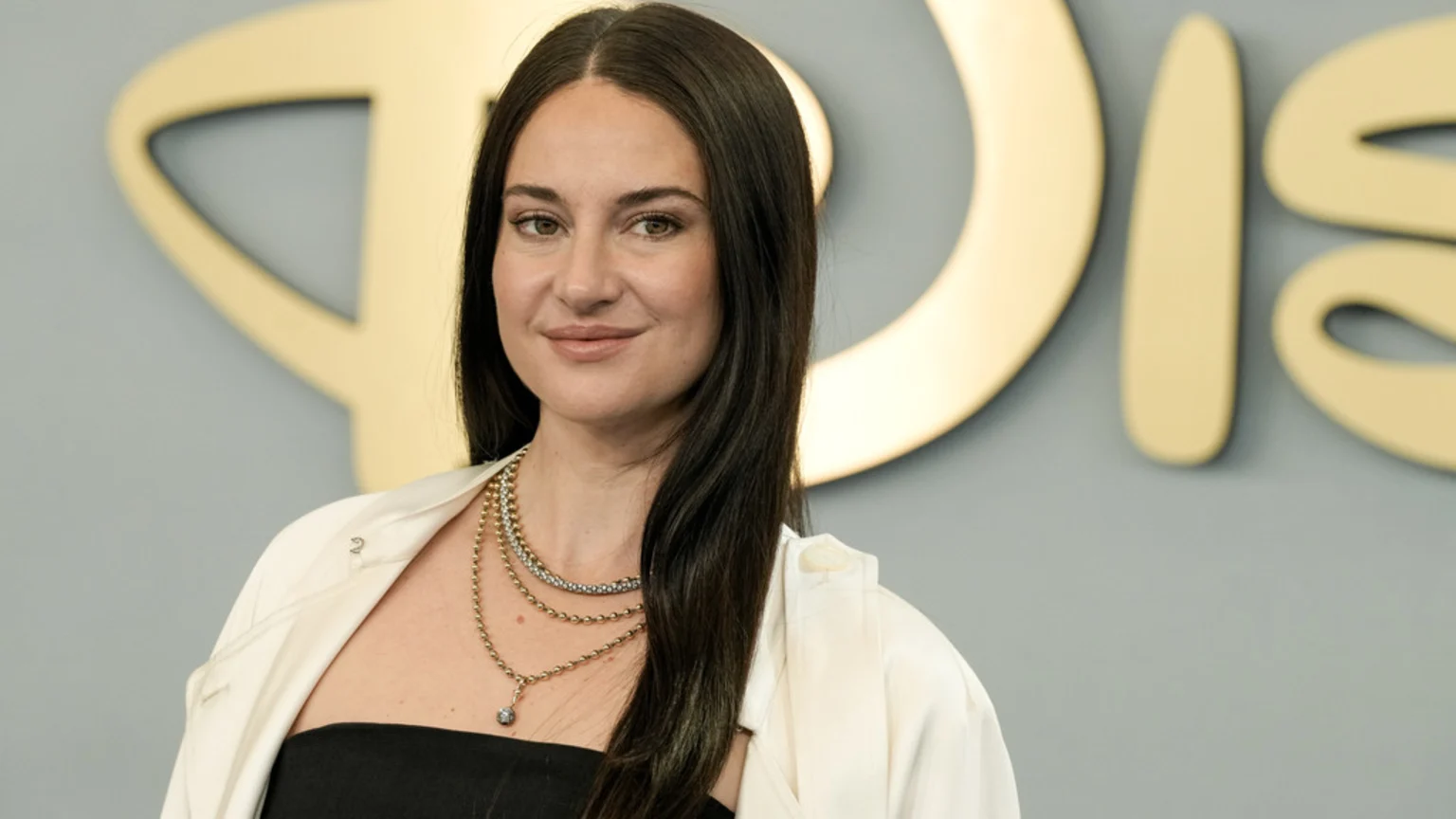 Shailene Woodley, with long dark hair, smiles slightly at the camera, wearing a black top, a white jacket draped over her shoulders, and layered gold necklaces, against a blurred gold and gray background.