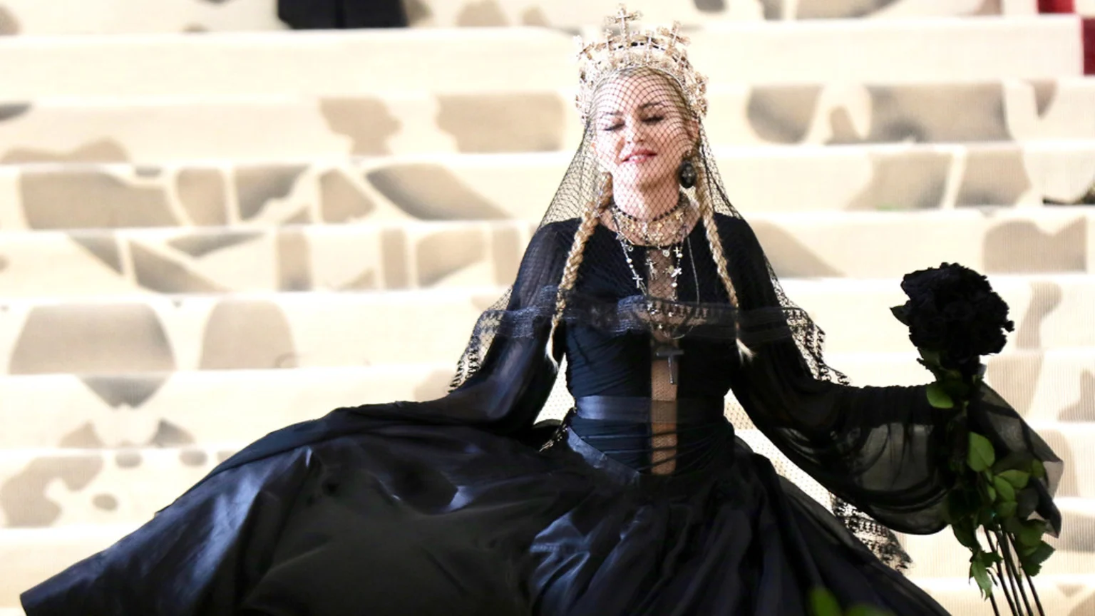 Madonna in a black gown, cross crown, and veil, holding black roses, smiles with closed eyes against a white patterned background.