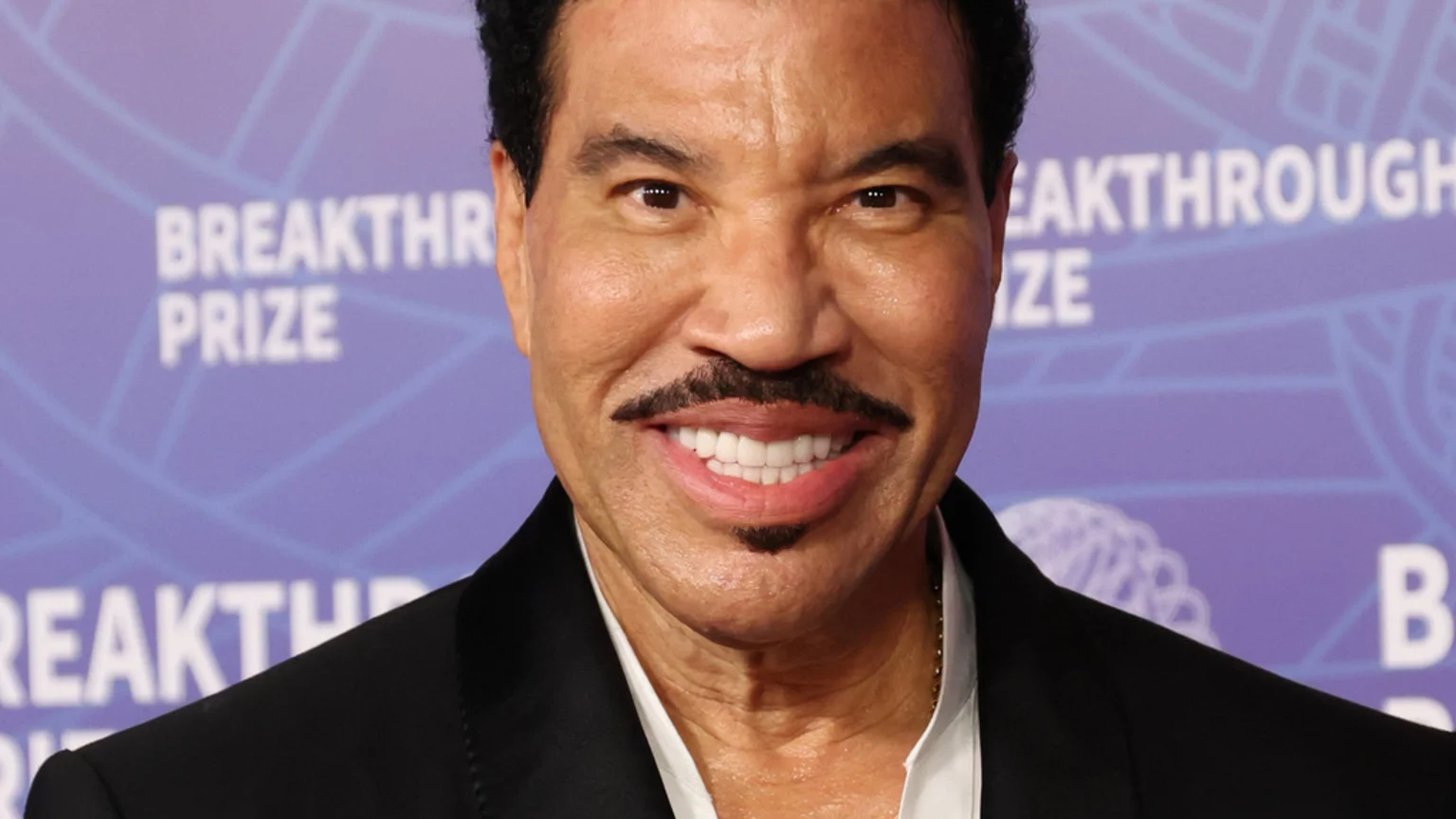 Lionel Richie smiles at the camera, wearing a black suit jacket and white shirt, with a mustache and goatee, against a blurred purple background with "BREAKTHROUGH PRIZE" text.