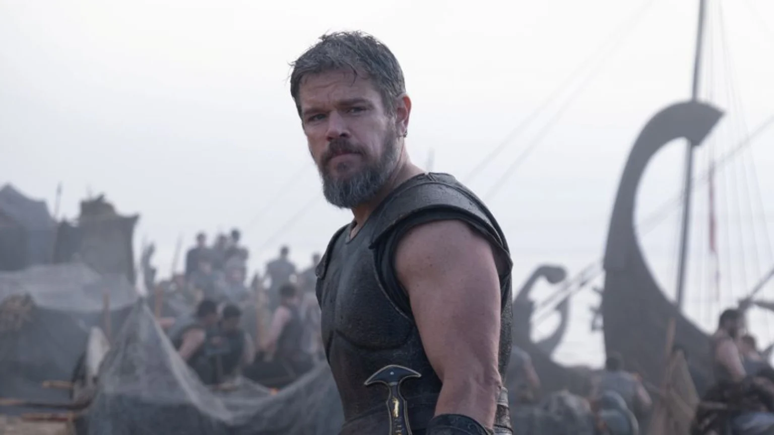 Matt Damon, a bearded man in dark armor, looks directly at the viewer, with a sword hilt visible at his side and blurred figures and ships in the hazy background.