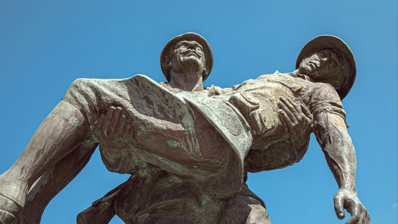 Bronze sculpture of two soldiers, one carrying the other, against a clear blue sky, seen from a low angle.