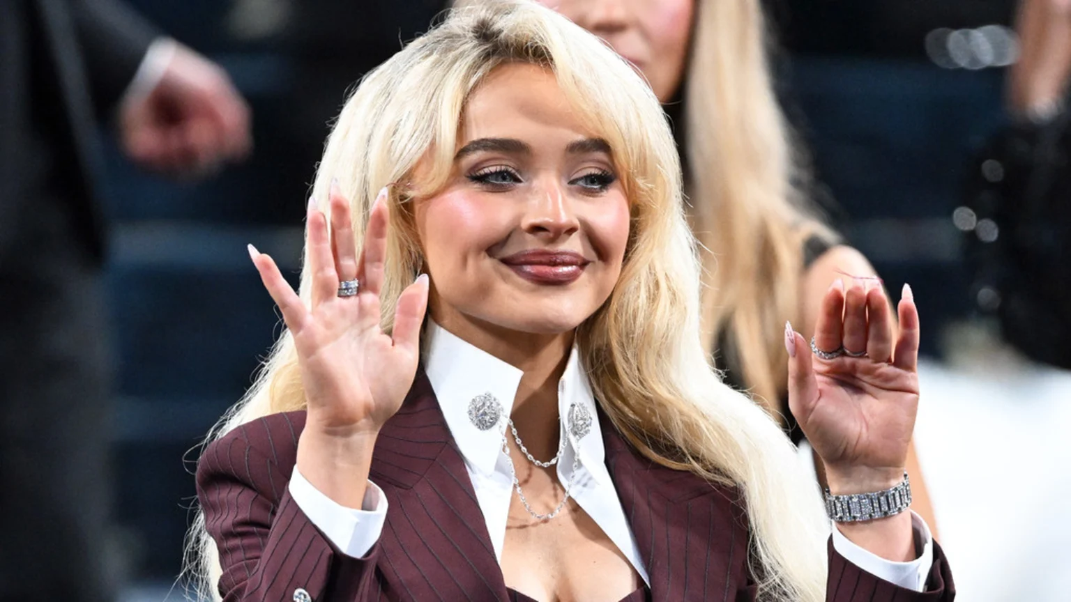 Sabrina Carpenter, a young woman with long blonde hair, smiles and raises both hands, wearing a pinstriped blazer, white shirt, and jewelry including rings and a diamond bracelet.