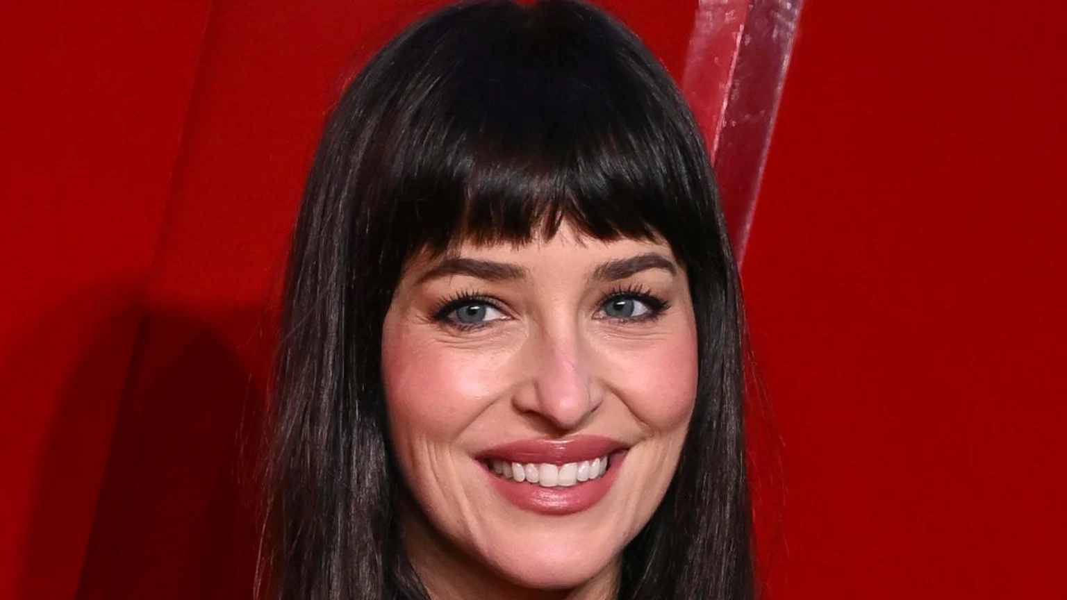 A woman with dark hair and bangs, blue eyes, and a wide smile, wearing makeup, against a solid red background.