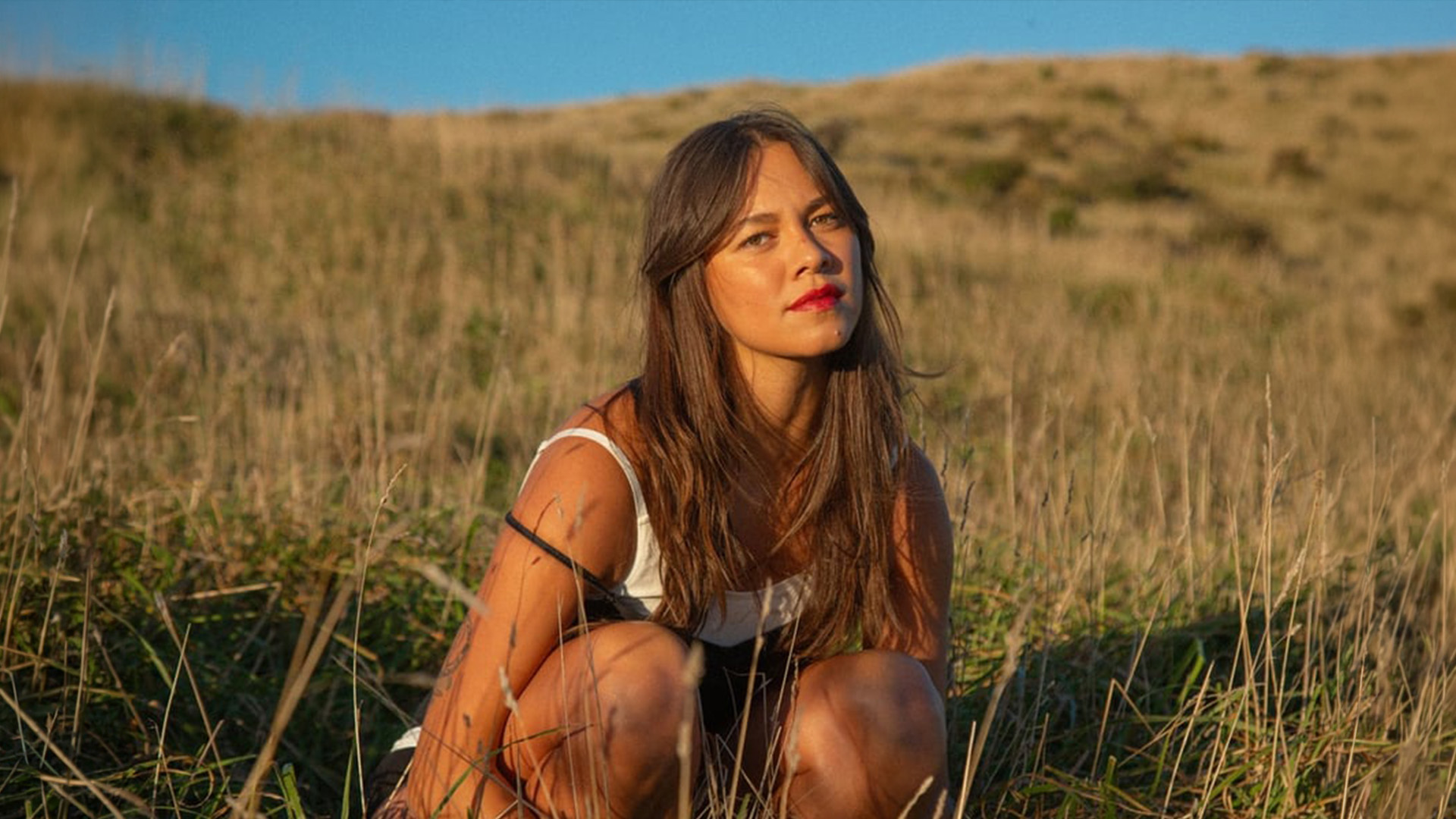 A woman with long brown hair and red lipstick crouches in a field of dry grass under a blue sky, looking towards the viewer.