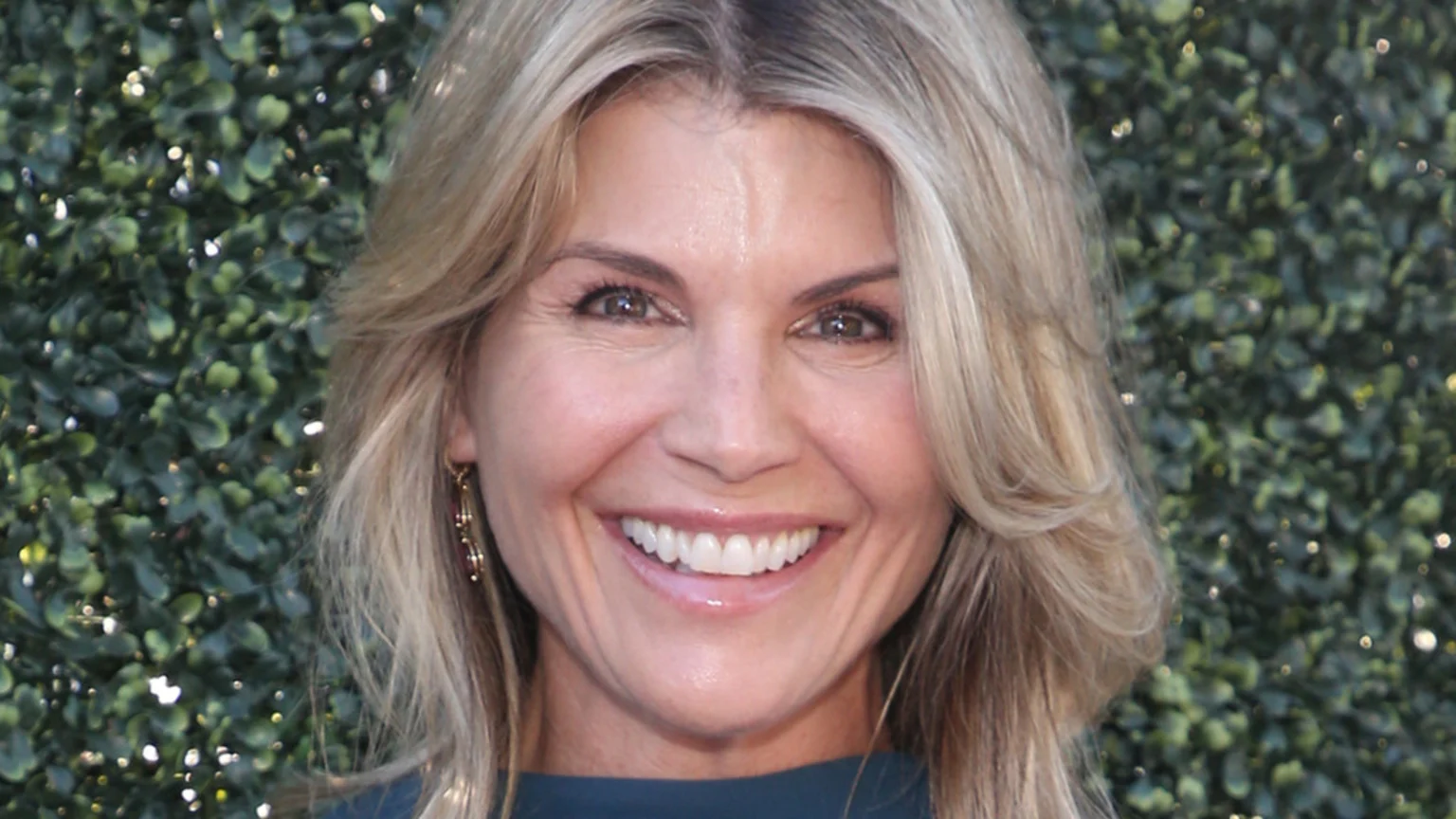 A smiling woman with blonde hair and brown eyes, wearing a dark top and earrings, poses against a blurred green leafy background.