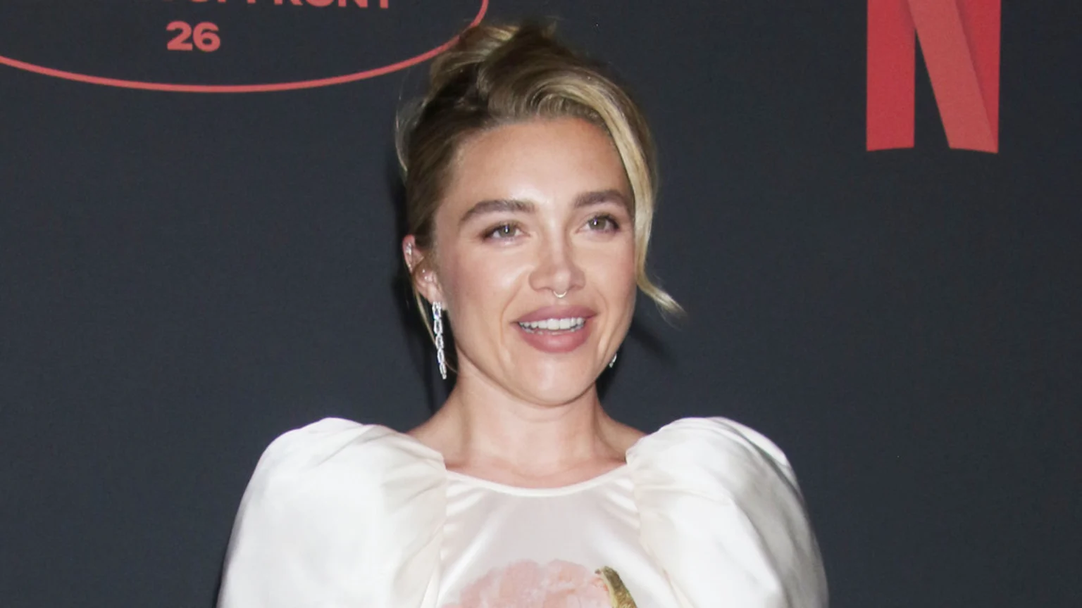 Florence Pugh smiles at the camera, wearing a light-colored dress with puffy sleeves, a septum piercing, and dangling earrings.