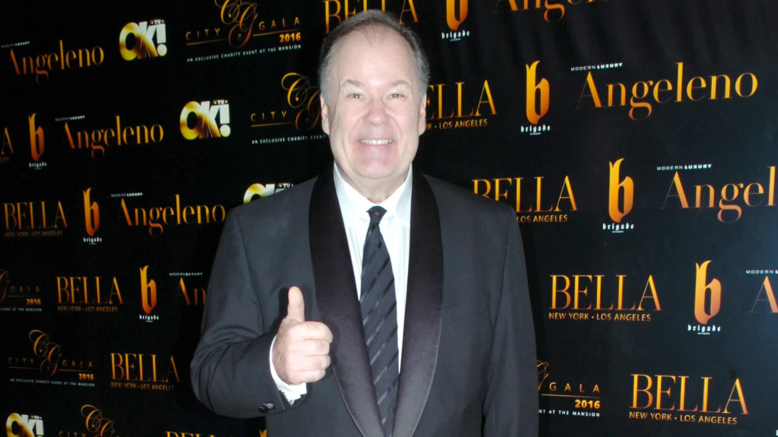 A smiling man in a black tuxedo gives a thumbs-up, standing against a black event backdrop featuring logos like "Angeleno," "OK! TV," and "BELLA New York - Los Angeles."
