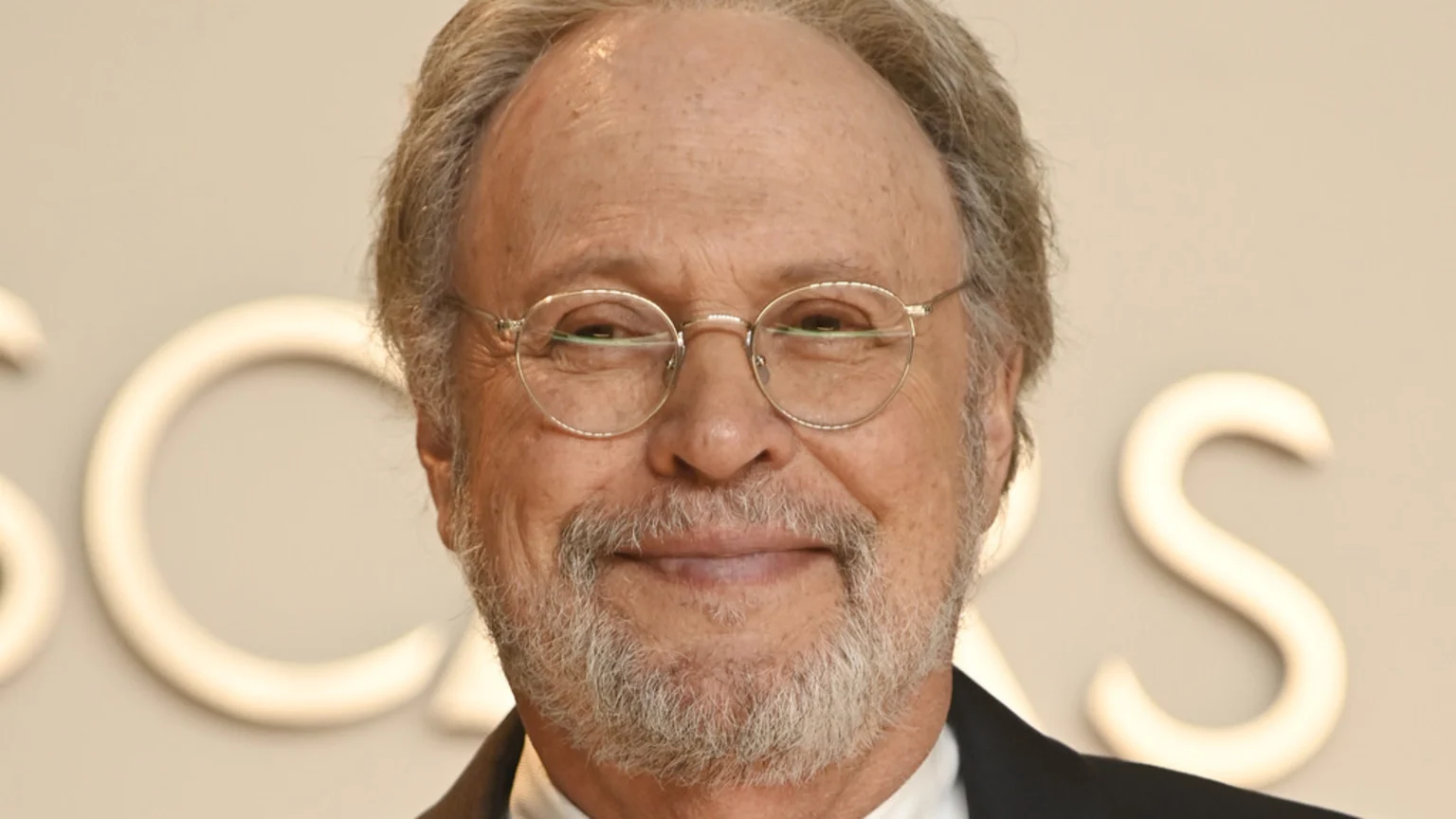 Close-up portrait of Billy Crystal smiling, wearing glasses and a grey beard, with blurred white letters like "OSCARS" in the background.