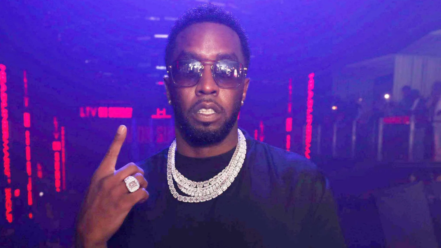 Sean "Diddy" Combs looks directly at the camera, wearing sunglasses, multiple diamond chains, and a ring, pointing his finger up in a dimly lit, purple-hued room with red lights.