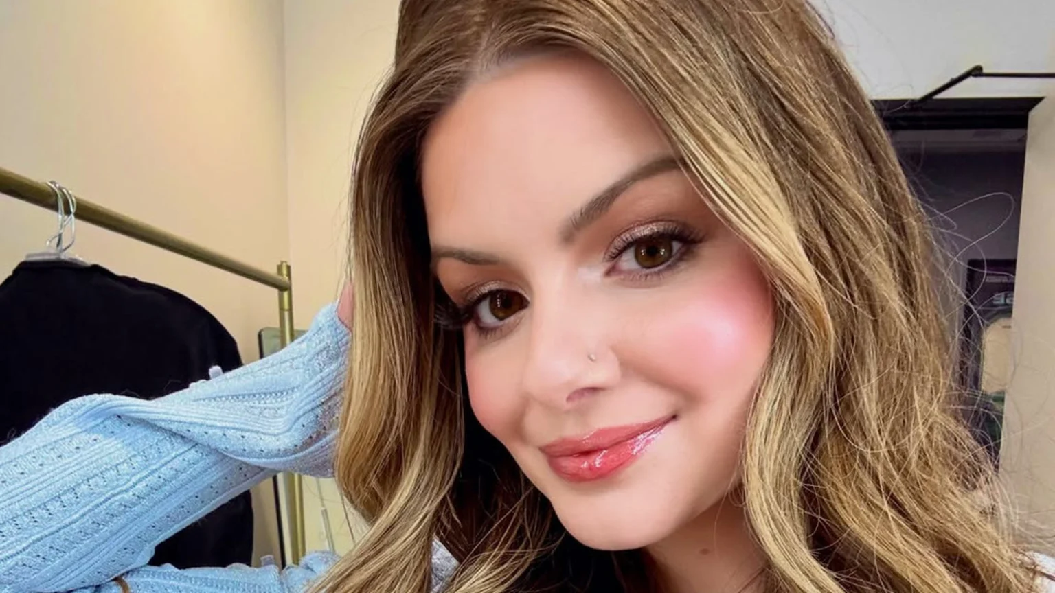 A close-up of a smiling young woman with long, wavy brown hair, wearing makeup with pink blush and glossy lips, a nose stud, and a light blue textured top.