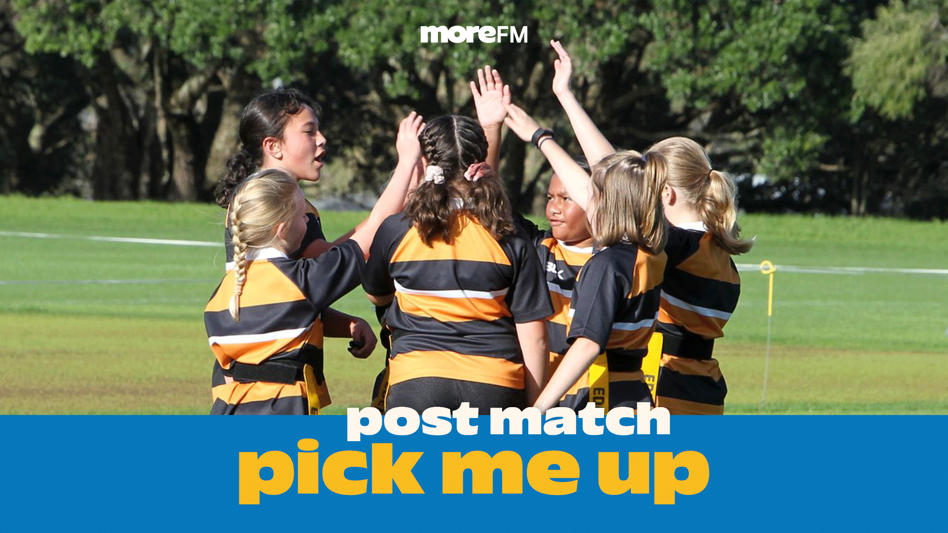 A group of young girls in black and orange striped uniforms huddle together on a grassy field, raising their hands in a collective gesture of teamwork and spirit.