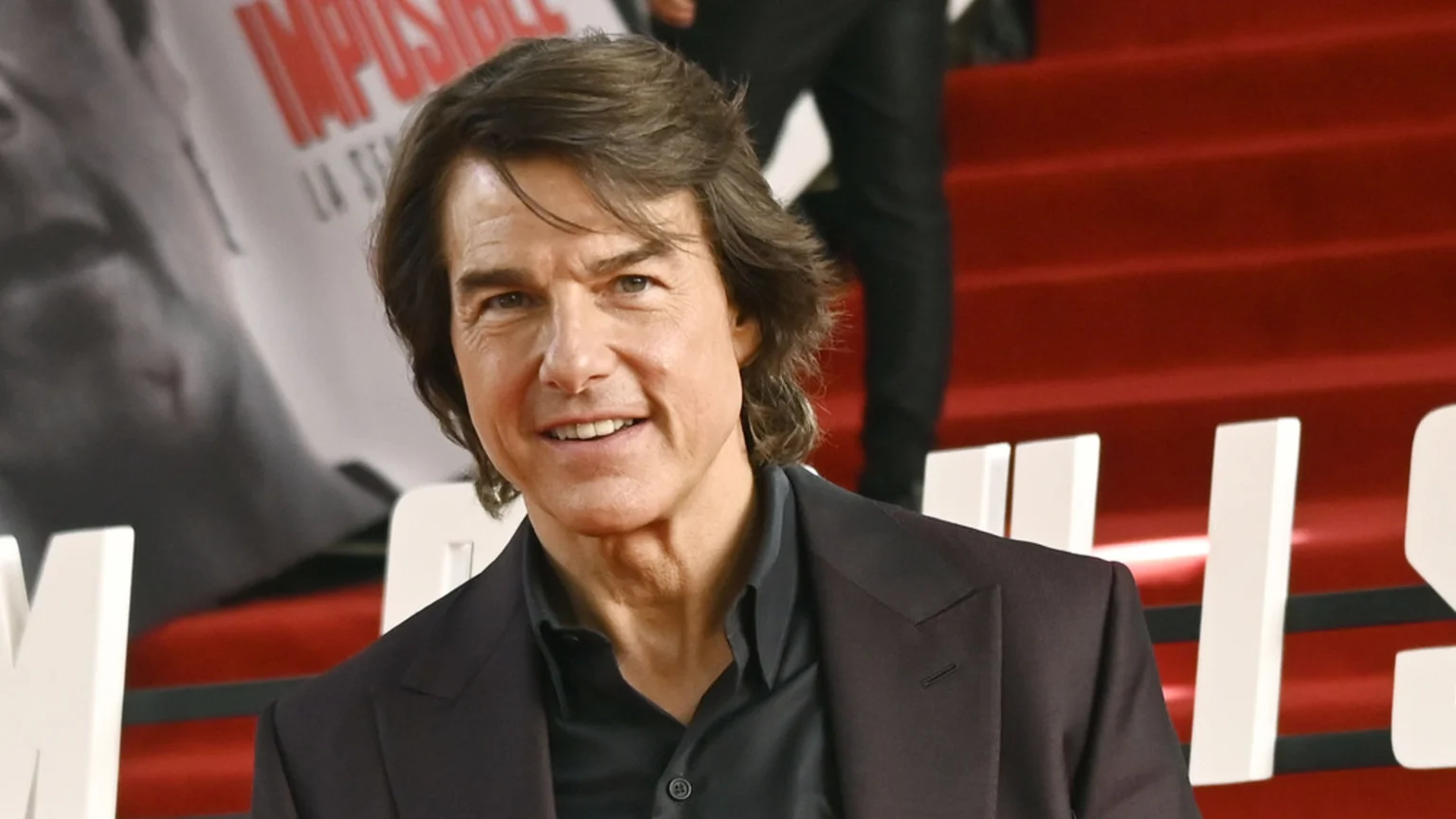 Tom Cruise smiles, wearing a dark suit jacket and shirt, standing in front of a red background and a banner with "IMPOSSIBLE" written on it.