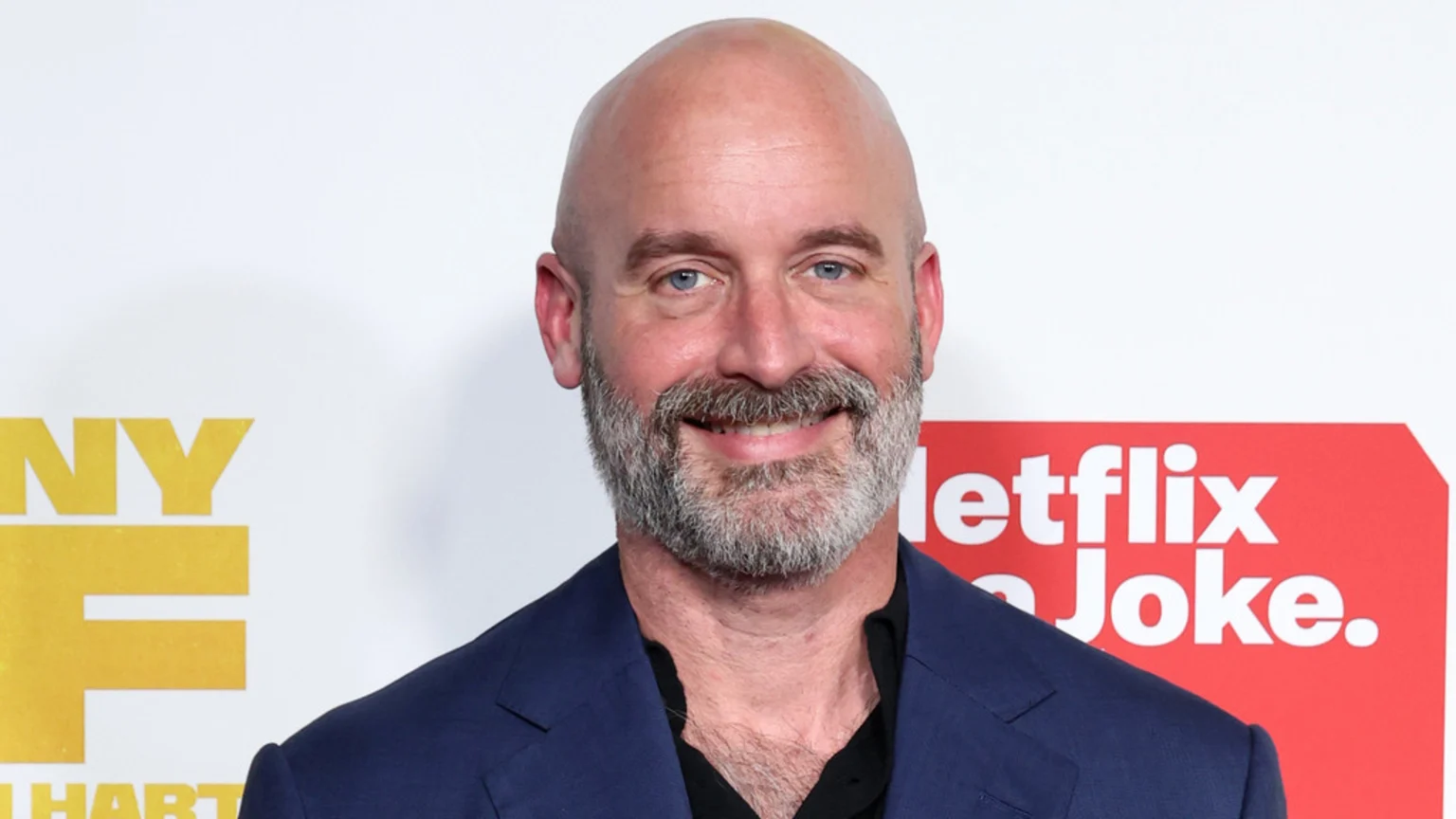 A smiling bald man with a gray beard and blue eyes, wearing a dark blue blazer and a black shirt, stands in front of a white wall with yellow and red logos.