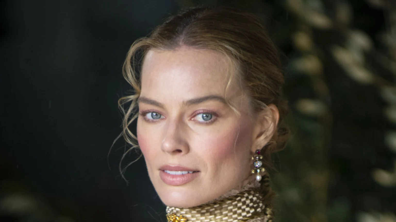 A close-up portrait of Margot Robbie with blonde hair styled in an updo, light blue eyes, and subtle makeup. She wears an elaborate patterned gold and brown high-collar garment and a detailed dangling earring featuring colorful gemstones and a white pearl. The image is set against a dark, softly lit, blurred studio background.