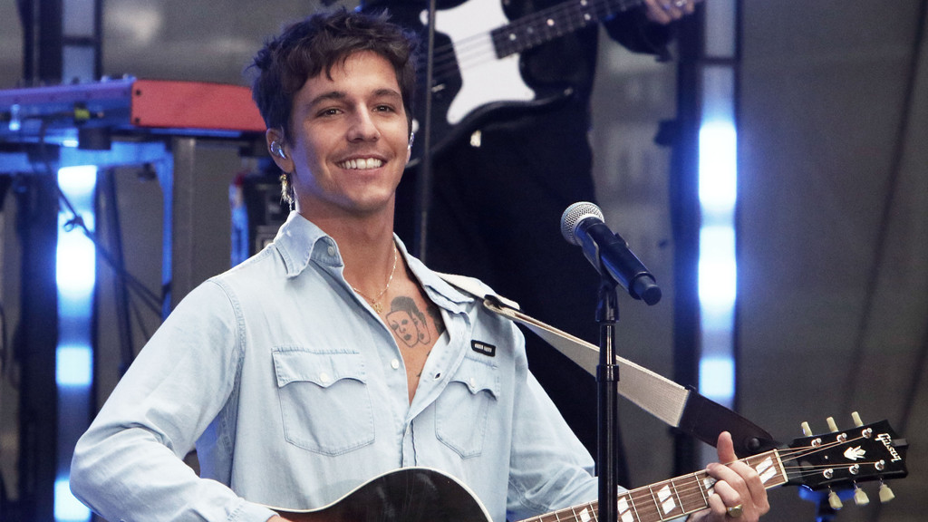 Singer Role Model smiles while playing an acoustic Gibson guitar, wearing a light blue denim shirt unbuttoned to show a chest tattoo and a gold chain, with a microphone nearby and a bass player in the background.