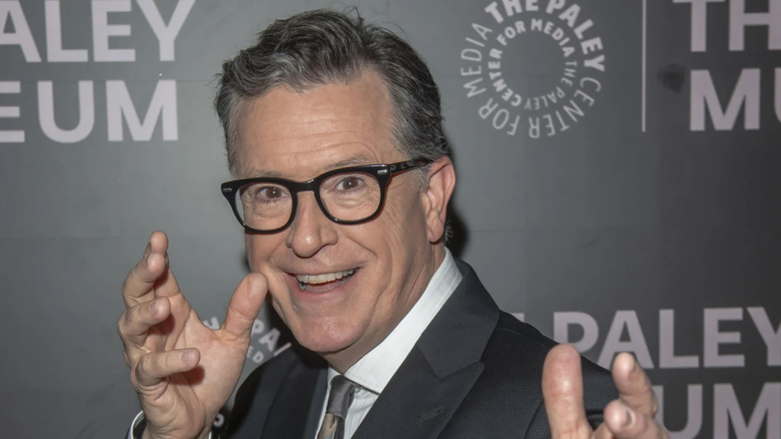 Stephen Colbert smiles widely at the camera, wearing a suit and glasses, with both hands gesturing in front of him against a grey background with blurred white text.