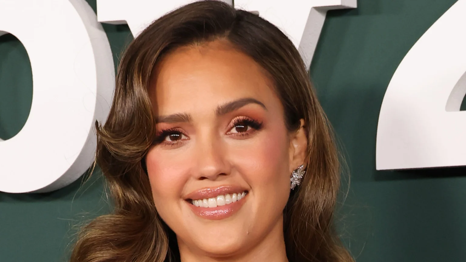 Smiling Jessica Alba in a close-up, with brown hair, warm-toned makeup, and a sparkling earring, against a dark green background with white letters.